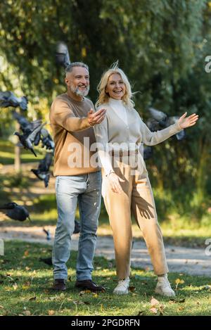 Die ganze Länge eines glücklichen Paares mittleren Alters, das neben fliegenden Tauben im Park steht, Stockbild Stockfoto