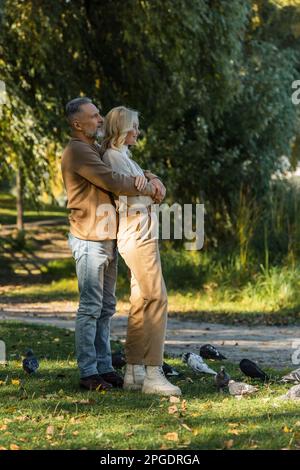 Die ganze Länge eines glücklichen Mannes mittleren Alters, der die blonde Frau umarmt, während er neben Tauben im Park steht, Stockbild Stockfoto