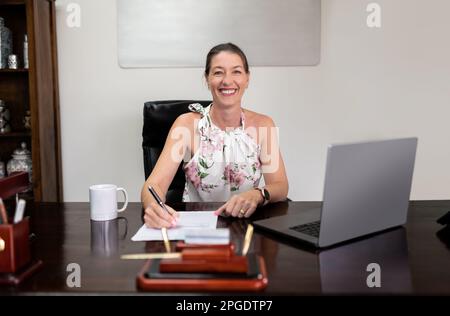 Eine glückliche Frau sitzt am Schreibtisch und schaut in die Kamera, während ihr Laptop vor ihr offen ist. Sie nutzt drahtlose Technologie für die Geschäftskommunikation Stockfoto