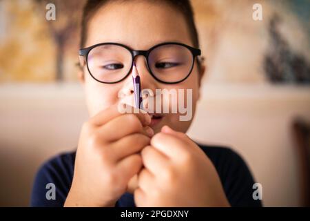 Ein schlauer, junger asiatischer Junge mit Brille schielt auf den Bleistift. Das Sehkrankheitsproblem. Stockfoto