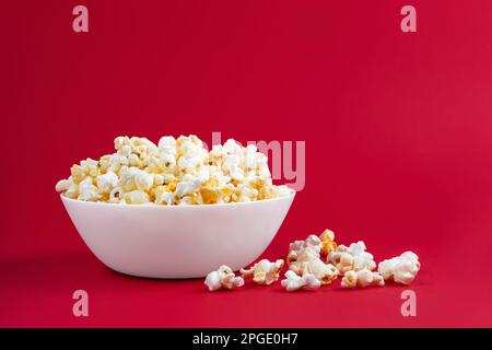 Verstreutes, leckeres Käse-Popcorn in weißer Schüssel auf rotem Hintergrund. Flach liegend, Draufsicht. Fast Food, Film, Kinokonzept Stockfoto