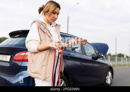 Eine Fahrerin hält ein Voltmeter vor dem Hintergrund eines kaputten Autos Stockfoto