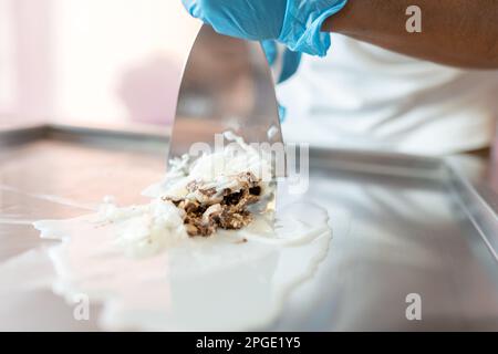 Ein Arbeiter verwendet Spatel, um Schokolade, gestreifte Kokosnuss und Milch auf einer Eiscreme-Maschine zu mischen Stockfoto