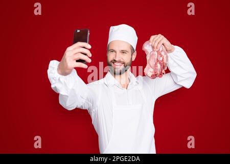 Porträt eines attraktiven, gutaussehenden Metzgers mit Bartstoppel-Selfie auf der Frontkamera des Smartphones, mit frischem Fleisch in der Hand, Blogger mit Video Stockfoto