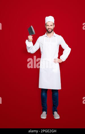 Ganzkörperporträt von positiv fröhlichem Metzger in Baskenmütze mit Hackbeil im Arm, der die Hand an der Taille hält, in die Kamera schaut, isoliert auf grau Stockfoto