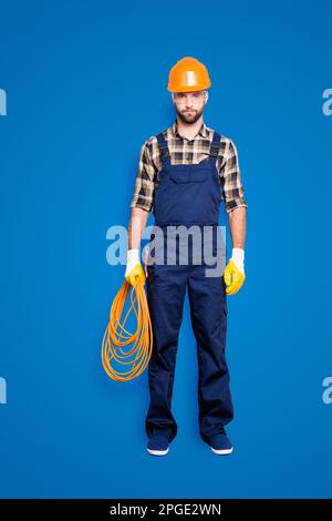 Vollkörperporträt in voller Größe, ein hübscher, strenger Elektriker mit Borsten im Overall, Hemd, gewalztes Seil in der Hand halten, in die Kamera schauen Stockfoto