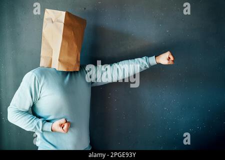 Seitenansicht eines Mannes mit einer Papiertüte am Kopf mit geballten Fäusten in Kampfposition, der Karate-Schläge auf grauem Hintergrund übt Stockfoto