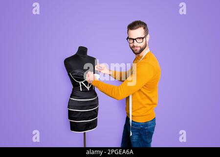 Portrait eines attraktiven, netten Schneiders in Pullover und Jeans mit Zentimeter am Hals, der Maße von Kleid an Schaufensterpuppe mit Nadeln sieht Stockfoto