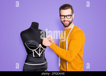 Portrait eines fröhlichen berühmten Schneiders im Pullover mit Zentimeter am Hals, der Messungen von eleganten Kleidern an Schaufensterpuppe mit Blick auf die Kamera durchführt Stockfoto