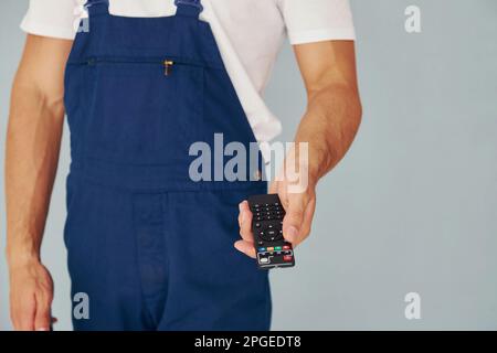 Nahaufnahme eines männlichen Arbeiters in blauer Uniform, der im Studio vor weißem Hintergrund steht und die Fernbedienung des Fernsehers hält Stockfoto