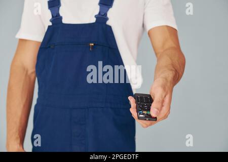 Nahaufnahme eines männlichen Arbeiters in blauer Uniform, der im Studio vor weißem Hintergrund steht und die Fernbedienung des Fernsehers hält Stockfoto