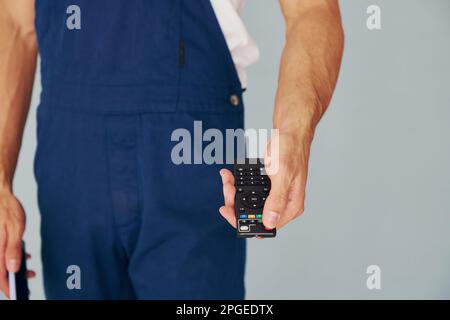 Nahaufnahme eines männlichen Arbeiters in blauer Uniform, der im Studio vor weißem Hintergrund steht und die Fernbedienung des Fernsehers hält Stockfoto