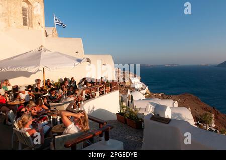 Happy Hour auf einer Panoramaterrasse am Meer im Sommer mit vielen Leuten, die an Tischen sitzen und den Sonnenuntergang in Oia, Griechenland, beobachten Stockfoto