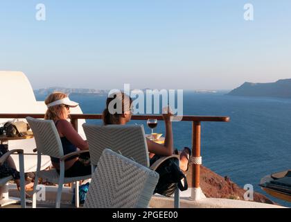 Dachbar mit Blick auf das Mittelmeer in Oia. Enge Freunde stoßen auf ihren griechischen Inselurlaub in einer schicken Terrassenbar am Meer mit Blick auf den Sonnenuntergang an Stockfoto