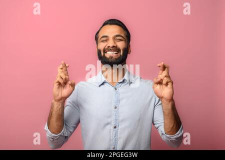 Wünsch mir Glück. Lächelnder junger indischer bärtiger Mann, der die Daumen gedrückt hält, die Augen geschlossen in der Hoffnung, ein multiethnischer Kerl will wirklich Glück haben, isoliert auf rosa Wand Stockfoto