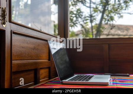 Laptop und Handy auf einem farbenfrohen Schreibtisch neben einem Fenster und Holzwänden Stockfoto