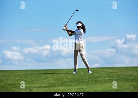 Minimales, durchgehendes Porträt einer sportlichen jungen Frau, die Golf auf einem grünen Feld gegen den Himmel spielt und einen schwingenden Golfschläger mit Präzision und Kopierbereich Stockfoto