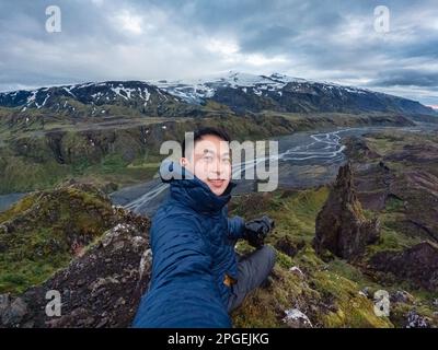 Junger männlicher asiatischer Wanderer, der an einem düsteren Sommertag in Thorsmork, Island, auf dem Gipfel mit dem Berg in den isländischen Highlands Selfie macht Stockfoto