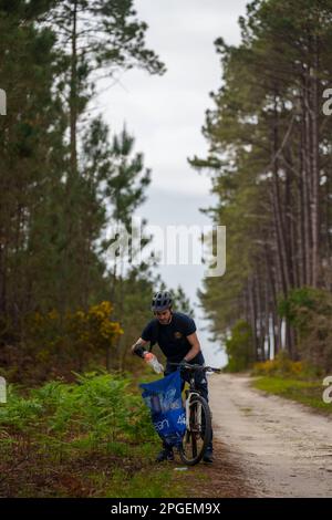Junger Mann, der Plastikflaschen und anderen Müll in einen Müllsack packt, während er auf einem Fahrrad durch den Wald pflückt Stockfoto