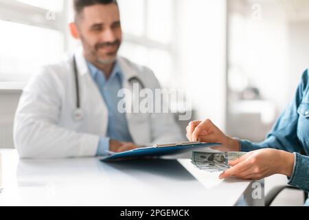 Weibliche Patientin, die Geld unter das Klemmbrett des Arztes legt, am Tisch in der Klinik sitzt, ein Mann, der es ansieht und lächelt Stockfoto