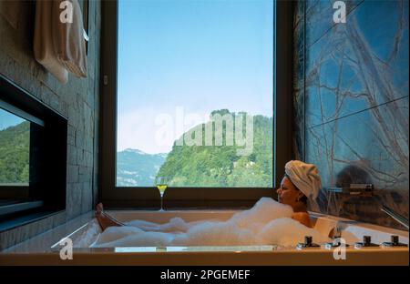 Frau liegt in einer Badewanne mit einem Glas Champagner und genießen den Fensterblick über den Berg an einem sonnigen Sommertag in Burgenstock, Nidwalden, Schweiz Stockfoto