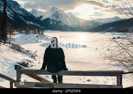 Rückansicht eines Reisenden, der im Winter im Jasper National Park, Alberta, Kanada, bei Sonnenaufgang über dem Medicine Lake sitzt und sich dort freut Stockfoto