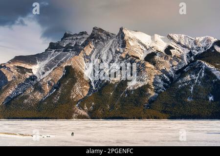 Schöner Blick auf den gefrorenen Lake Minnewanka mit felsigen Bergen im Winter am Abend im Banff National Park, Alberta, Kanada Stockfoto