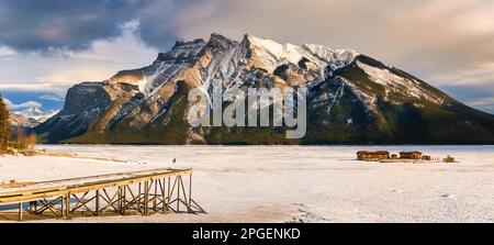 Panoramablick auf den schönen gefrorenen Lake Minnewanka mit felsigen Bergen im Winter am Abend im Banff National Park, Alberta, Kanada Stockfoto