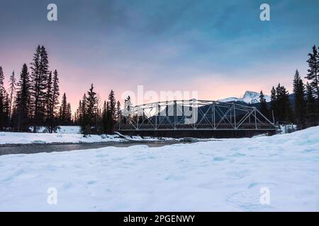 Im Banff National Park, Alberta, Kanada, geht die Sonne über verschneiten Felsbrocken auf und die Brücke überquert den Fluss im Kiefernwald Stockfoto