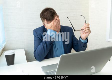 Müder oder erschöpfter Mann, der sein Gesicht mit Händen vor dem Computer im Büro schließt. Ein Geschäftsmann bedeckt sein Gesicht und seine Augen mit seinen Händen. Facepa Stockfoto