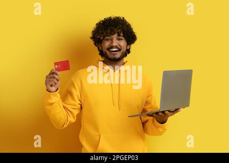 Fröhlicher Ost-Typ, der Laptop und Bankkarte zeigt Stockfoto
