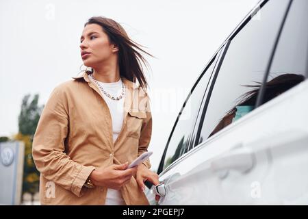 Er steht draußen und hält das Telefon. Modische, wunderschöne junge Frau und ihr modernes Auto Stockfoto
