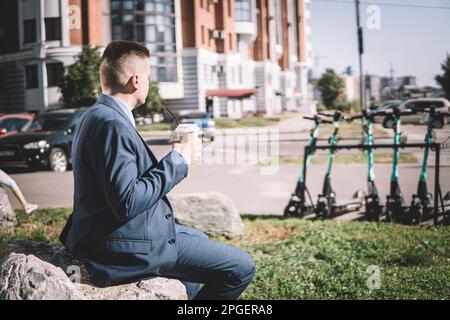 Junger Geschäftsmann im Business-Anzug, während er einen elektrischen Roller anschaut, der Kaffee im Hintergrund der Morgenstadt trinkt Stockfoto