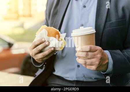 Geschäftsmann frühstückt. Fastfood, Junkfood in der Hand eines jungen Geschäftsmannes im Anzug. Stockfoto
