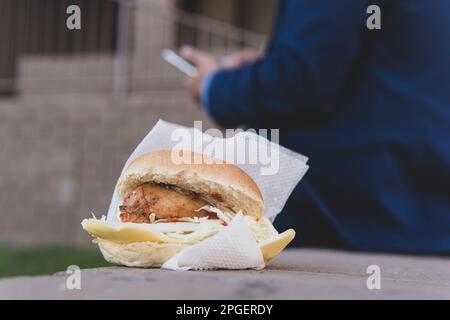 Hühnchen-Sandwich auf einer Bank auf der Straße. Das Konzept des Junkfood in der Mittagspause. Geschäftsmann isst ungesunde kalorienreiche Lebensmittel auf der Straße Stockfoto