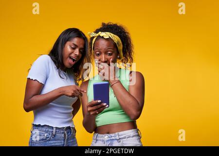 Porträt junge, überraschte schwarze Mädchen, die auf das Handy zeigen und schauen. Menschen isoliert auf Gelb. Stockfoto