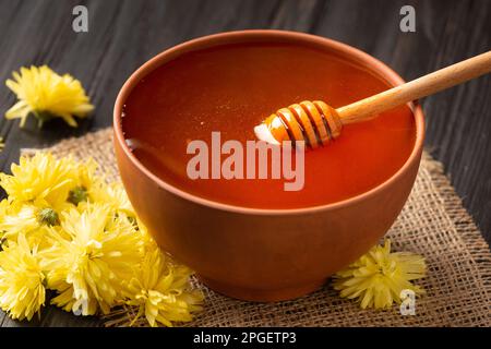 Honig in einer Tonschüssel, Dipper und gelbe Blumen auf Textilhintergrund. Ein Honigstock liegt in einer Tonschüssel mit Honignaht. Stockfoto