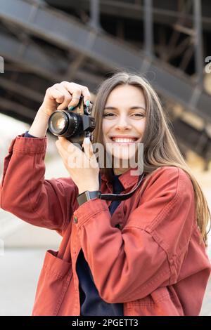 Nahaufnahme einer fröhlichen jungen Frau, die eine alte Kamera hält, lächelt und draußen in die Kamera schaut Stockfoto