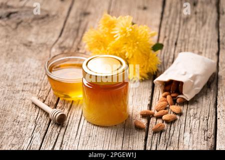 Honig in einem geschlossenen Glasgefäß und einer Schüssel auf Holzhintergrund. Zusammensetzung von Honiggläsern, Dipper, Nüssen im Sack und gelben Blumen. Gesunde Lebensmittel. Stockfoto