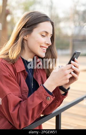 Porträt einer positiven jungen Frau, die mit ihrem Handy chattet, mit einem großen Lächeln im Gesicht und einem wunderschönen braunen Haar draußen Stockfoto