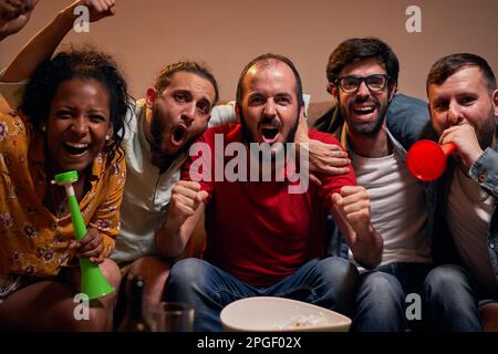 Fußballfans entspannen sich auf dem Sofa und sehen sich ein Sportspiel zu Hause an, wenn sie in die Kamera schauen Stockfoto