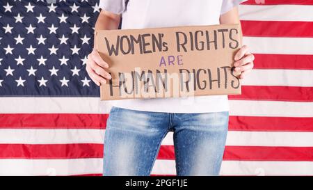 Junge Frau Protestierenden hält Pappe mit Womens Rights are Human Rights Zeichen gegen USA Flagge auf Hintergrund. Feministische Macht. Chancengleichheit für Frauen Stockfoto