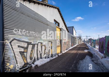 Industriehallen hinter einem mit Graffiti bedeckten Welleisenzaun im Industriegebiet Kyläsaari in Helsinki, Finnland Stockfoto