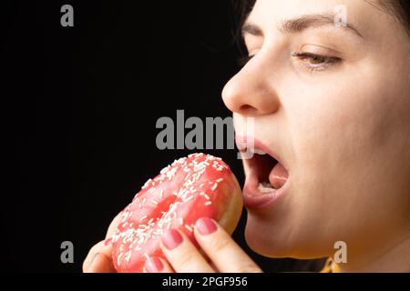 Eine Frau beißt einen großen roten Donut, einen schwarzen Hintergrund, ein Ort für Text. Gluttonie, überfressen und zuckersüchtig. Stockfoto