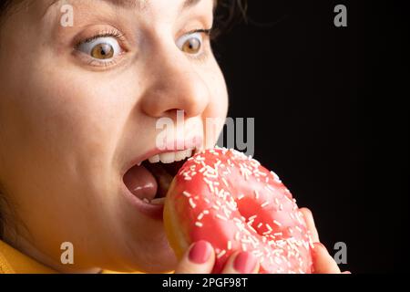 Eine Frau beißt einen großen roten Donut, einen schwarzen Hintergrund, ein Ort für Text. Gluttonie, überfressen und zuckersüchtig. Stockfoto