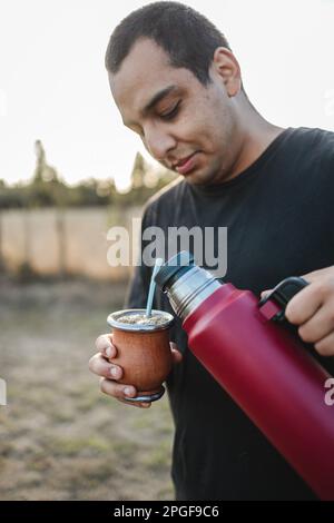 Junger lateiner, der Yerba-Mate trinkt, heißes Wasser aus einer roten Thermoskanne serviert, auf dem Land. Stockfoto