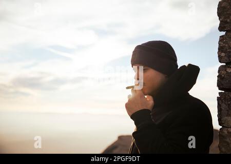 Ein Junge, der während des Sonnenuntergangs einen Marihuana-Joint auf einem Berg raucht Stockfoto