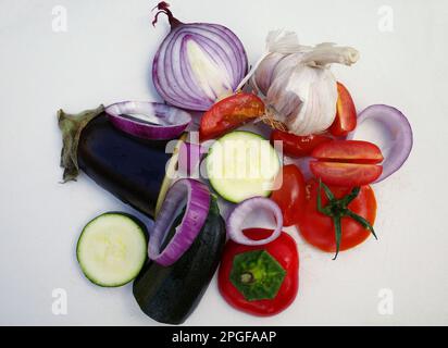 Gemischtes Gemüse für ein mediterranes Gericht auf einem weißen Schneidebrett. Rote Zwiebeln, Zucchini, Aubergine, Knoblauch, Tomaten und Paprika sind auf dem Teller Stockfoto