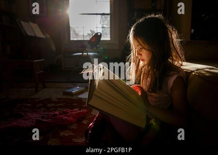 Ein kleines Kind sitzt im hellen Sonnenlicht und liest ein großes Buch Stockfoto