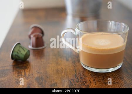 Tasse schaumiger Kaffee mit Milch neben einigen Kapseln auf dem Tisch Stockfoto